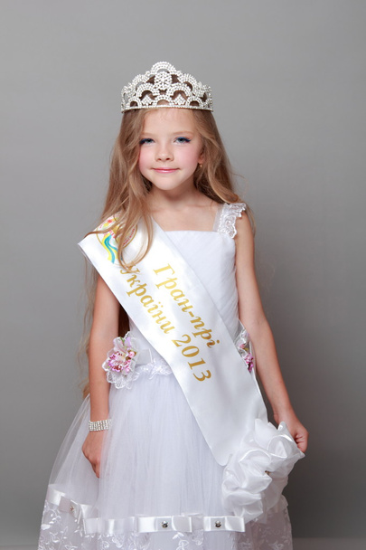 Cute little girl wearing a crown and a white dress cute smiling and posing for the camera on a gray background - 写真・画像