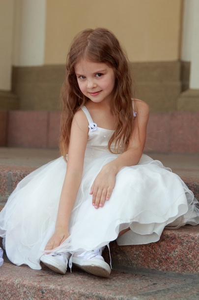 Smiling cheerful little girl in a beautiful white ball gown and sneakers sitting on the stairs to the outdoors - Photo, image