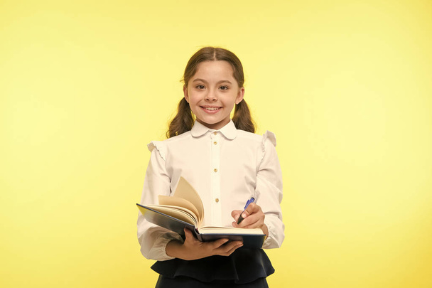 Write note to remember. Child school uniform smart kid happy make note. Girl happy face make note about idea yellow background. Child girl happy school uniform clothes holds book ready write note - Foto, Imagen
