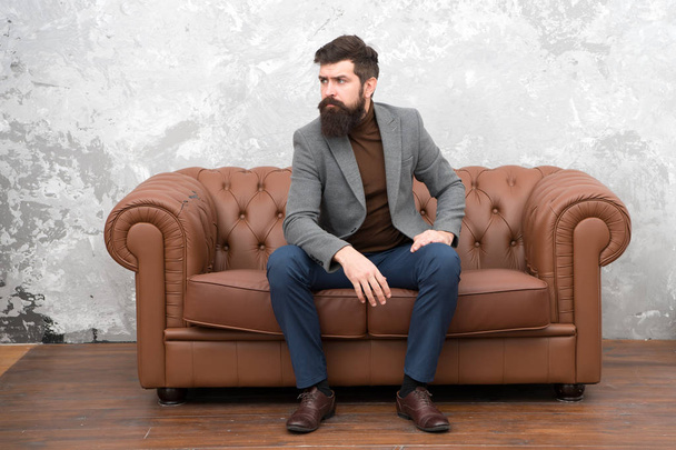 Realtor and rental service. Rent apartment. Bearded man with confident face sit leather couch. Loft interior apartment. Businessman realtor work. Furniture shop. Hipster realtor loft style apartment - Foto, Imagem