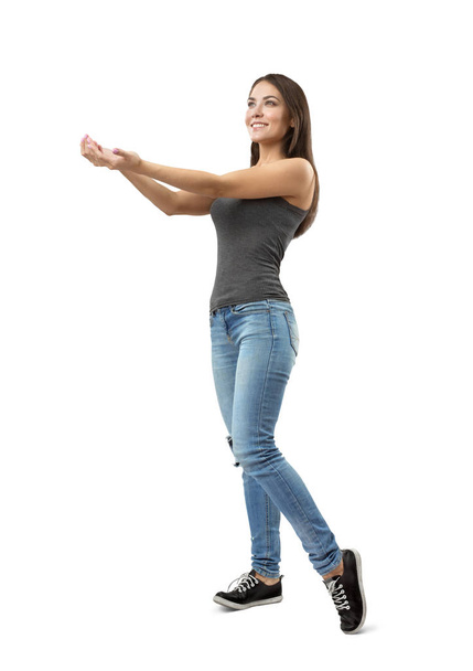 Young fit smiling woman in gray sleeveless top and blue jeans standing in half-turn holding out her hands with palms facing up on white background. - Фото, изображение