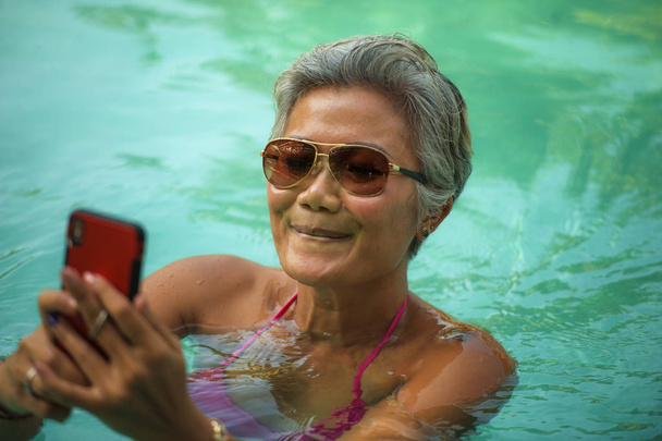 attractive and happy middle aged Asian Indonesian woman in bikini swimming at tropical luxury resort pool taking selfie portrait with mobile phone enjoying Summer holiday - Photo, Image