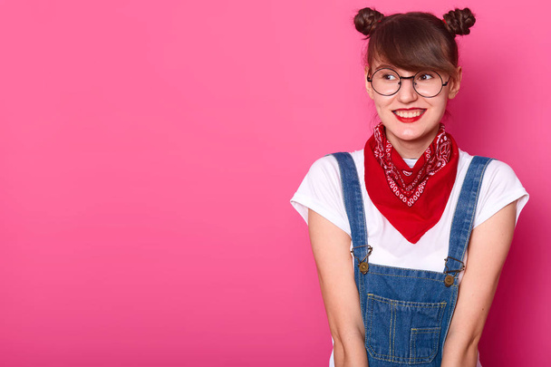 Young smiling sweet girl looking aside, wearing casual white t shirt, jeans overalls, red bandana and round trendy spectacles, being in high spirits, having bunches. Copy space for advertisement. - Фото, изображение