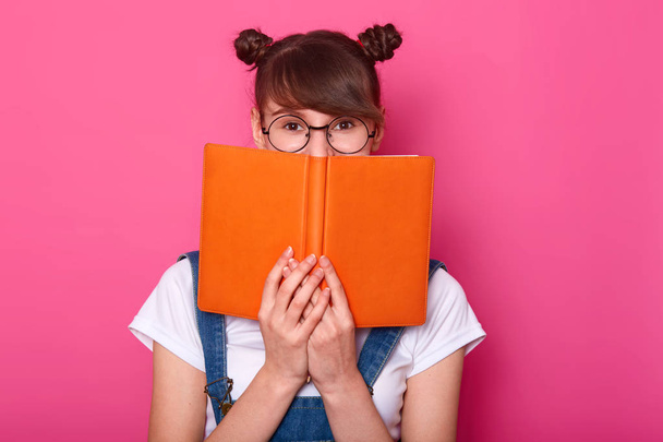 Happy pensive girl standing isolated over pink background in studio, holding orange notebook, covering half of face, looking attentively at camera, having bunches, wearing casual trendy clothes. - Fotó, kép