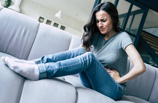 Female pain. Woman with pain in stomach is sitting on couch at home.  - Foto, Bild