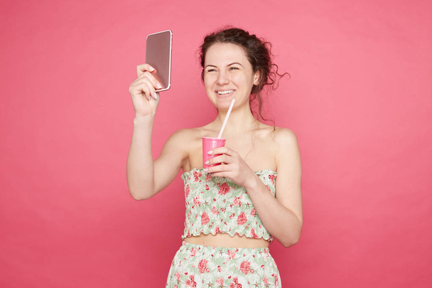 Vrolijke Kaukasische dame spreekt oprechte gevoelens en emoties uit, maakt selfie voor het delen van foto in sociaal netwerk, houdt Roze plastic glas met stro, draagt zomer vrouwelijke outfit, modellen in Studio. - Foto, afbeelding