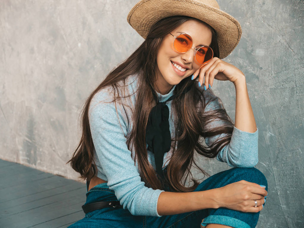 Portrait of Young beautiful smiling woman looking at camera. Trendy girl in casual summer clothes and hat. Funny and positive female posing near gray wall in studio in sunglasses. Sitting on the floor - Фото, изображение