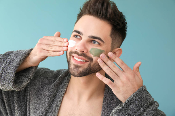 Handsome man applying clay mask on his face against color background - Foto, imagen