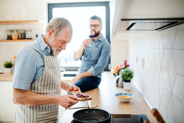 An adult hipster son and senior father indoors in kitchen at home, cooking. - 写真・画像