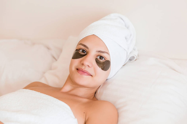 Beautiful young woman with under eye patches in bathrobe lying in bed. Happy girl taking care of herself. Beauty skincare and wellness morning concept - Zdjęcie, obraz