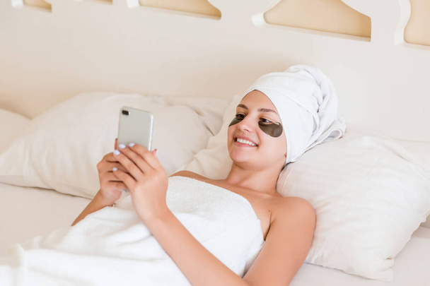 Gyönyörű fiatal nő szem alatti foltok és mobil telefon használata fürdőköpenyben feküdt az ágyban. Boldog lány vigyáz magára. Szépség bőrápoló és wellness reggel koncepció - Fotó, kép