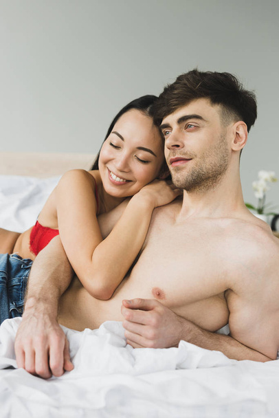 happy asian woman embracing pensive shirtless boyfriend in bedroom - Fotoğraf, Görsel