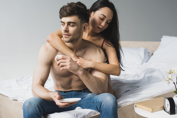 pretty asian woman hugging handsome boyfriend sitting on bed with cup of coffee - Photo, image
