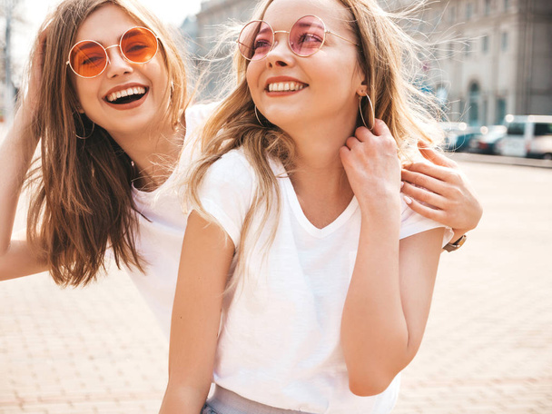 Portrait of two young beautiful blond smiling hipster girls in trendy summer white t-shirt clothes. Sexy carefree women posing on street background. Positive models having fun in sunglasses.Hugging - Foto, afbeelding