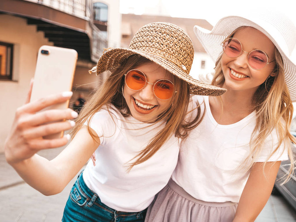 Two young smiling hipster blond women in summer white t-shirt clothes. Girls taking selfie self portrait photos on smartphone.Models posing on street background.Female showing positive face emotions - Photo, image