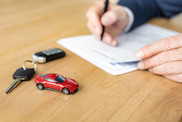selective focus of red toy car near car key and car dealer holding pen near contract  - 写真・画像