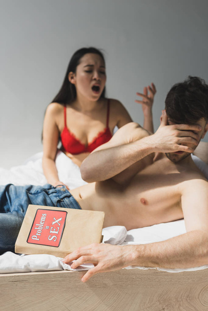 selective focus on angry asian woman in red lingerie screaming at boyfriend lying on bed and holding problems in sex book - Photo, Image