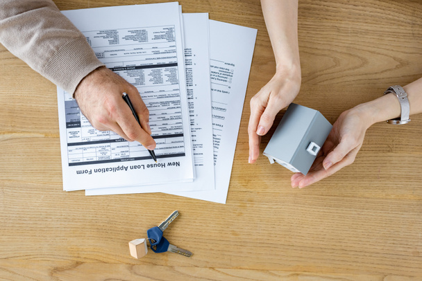 Ansicht von oben: Mann hält Stift in der Nähe von neuem Antragsformular für Hauskredite und Frau mit Hausmodell in der Hand  - Foto, Bild