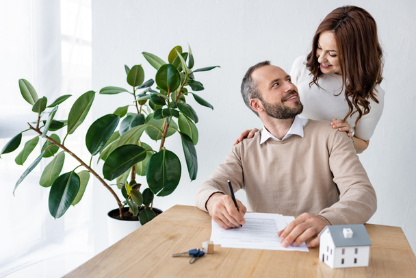 επιλεκτική εστίαση του άντρα με γένια κοιτάζοντας ευτυχισμένη γυναίκα κοντά στο μοντέλο του σπιτιού και τα κλειδιά στο τραπέζι  - Φωτογραφία, εικόνα