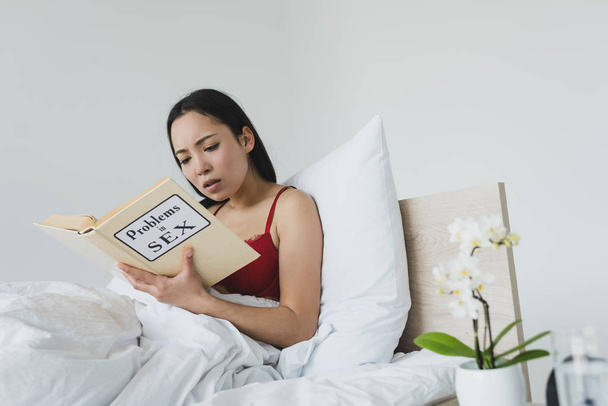 surprised asian woman reading problems in sex book while lying in bed - Фото, зображення