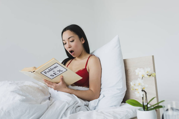 geschokt Aziatische vrouw lezen problemen in seks boek terwijl liggend in bed - Foto, afbeelding