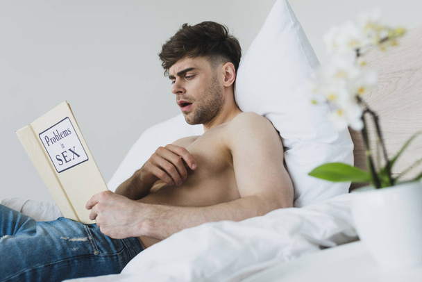 σοκαρισμένος όμορφος άνθρωπος διαβάζοντας προβλήματα στο σεξ βιβλίο, ενώ κείτεται σε λευκά κλινοσκεπάσματα - Φωτογραφία, εικόνα