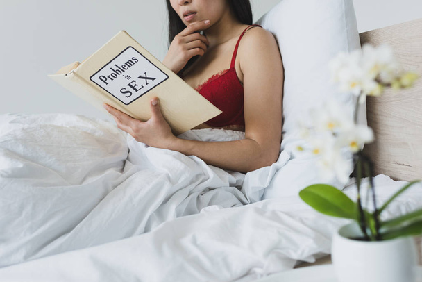 young woman in red lingerie reading problems in sex book while lying in bed - Photo, Image