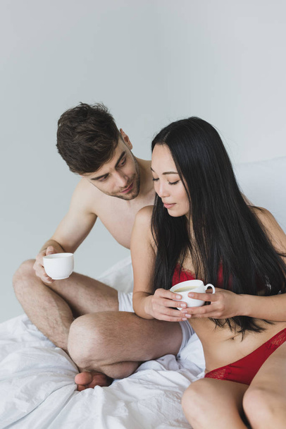heureux couple interracial assis sur le lit avec les jambes croisées et tenant des tasses à café
  - Photo, image