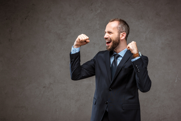 happy bearded businessman in suit gesturing while celebrating on grey  - 写真・画像