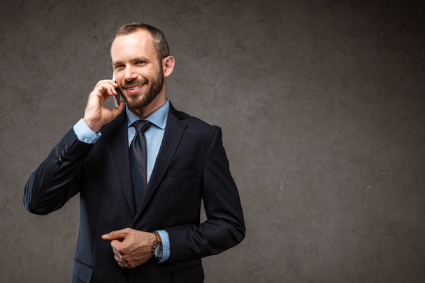 χαρούμενος επιχειρηματίας μιλάει στο smartphone και χαμογελάει σε γκρι  - Φωτογραφία, εικόνα