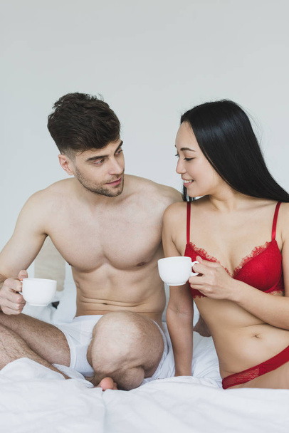 cheerful interracial couple sitting on white bedding and holding coffee cups  - Fotó, kép