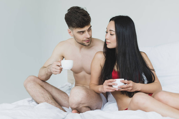 sorridente coppia interrazziale seduta su biancheria da letto bianca e in possesso di tazze di caffè
  - Foto, immagini