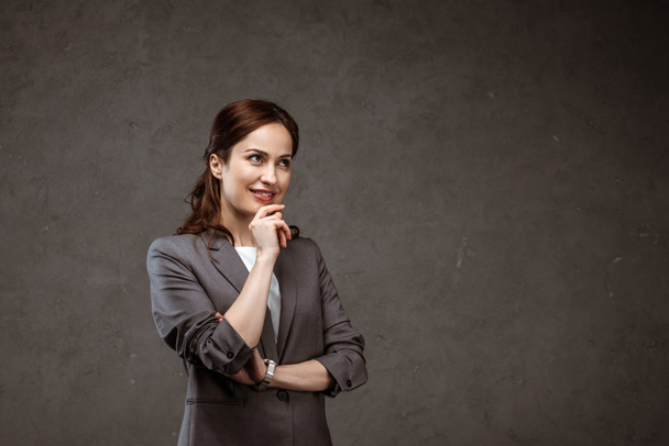 pensive brunette businesswoman smiling while standing on grey  - Photo, Image