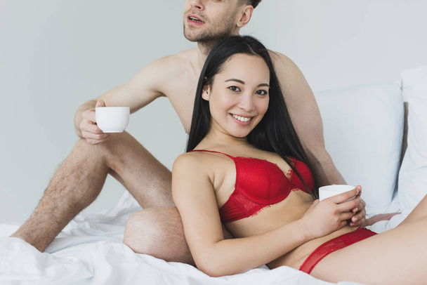 partial view of shirtless man holding coffee cup while sitting on white bedding near smiling  asian girlfriend - Foto, Imagem