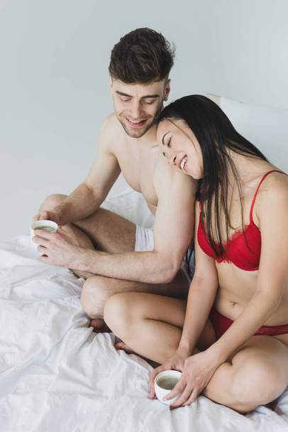 cheerful loving interracial couple sitting on bed with crossed legs and holding coffee cups  - Фото, изображение