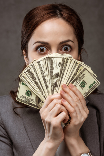 šokovaná brunetka zakrývající tvář dolarovou bankovkami na šedé  - Fotografie, Obrázek