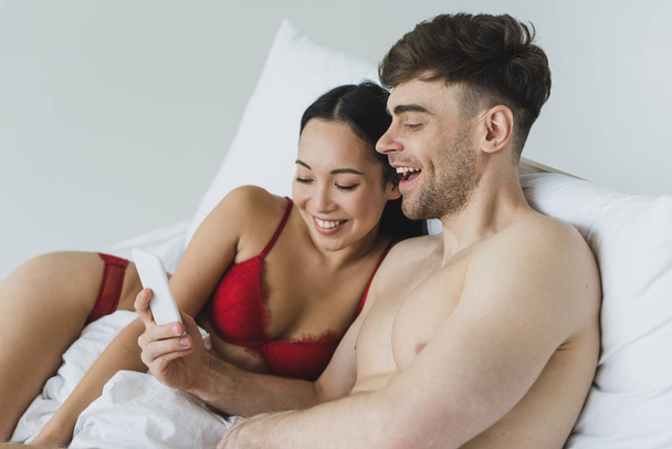 cheerful interracial couple in underwear lying in bed and using smartphone together - Foto, Imagem