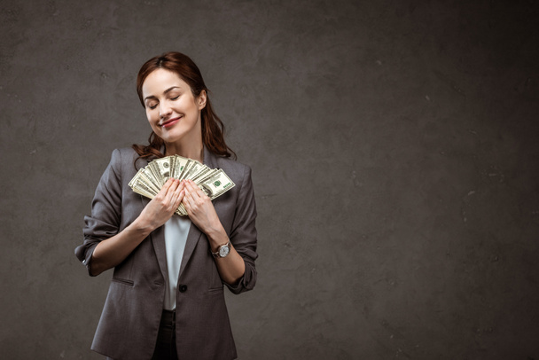 gelukkige zakenvrouw staande met gesloten ogen en het houden van dollar bankbiljetten op grijs  - Foto, afbeelding