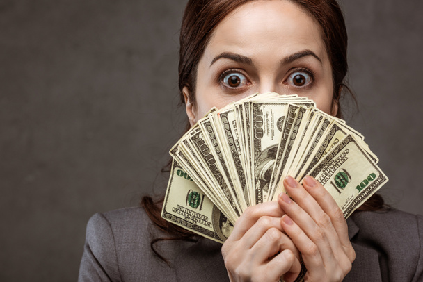 surprised brunette businesswoman covering face with dollar banknotes on grey  - Photo, Image
