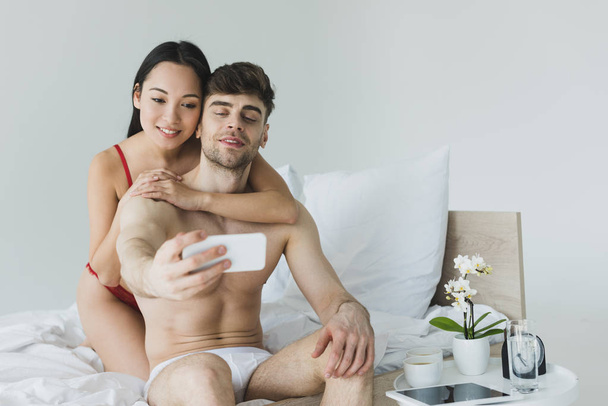 felice coppia interrazziale seduta su biancheria da letto bianca in biancheria intima e scattare selfie con smartphone
 - Foto, immagini
