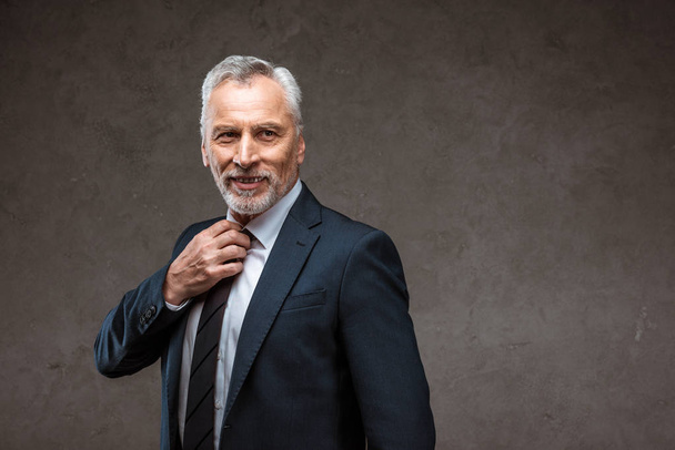 cheerful bearded businessman touching tie while smiling on grey  - Foto, Imagem