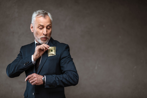 bearded businessman in suit putting dollar banknote in pocket on grey  - Фото, изображение