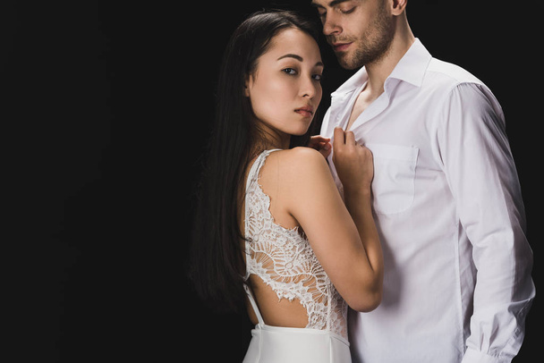 attractive asian woman in white lingerie standing near boyfriend and looking at camera isolated on black - Fotoğraf, Görsel