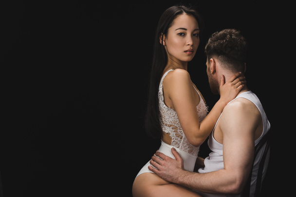 sexy asian woman in white lingerie sitting on knees of boyfriend and looking at camera isolated on black - Φωτογραφία, εικόνα