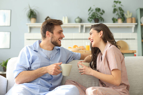 Beautiful young couple in pajamas is looking at each other and smiling on a sofa in the living room. - Foto, Imagen