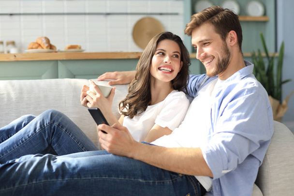 Jovem casal assistindo conteúdo online em um telefone inteligente sentado em um sofá em casa na sala de estar. - Foto, Imagem