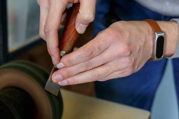 Το κλείσιμο του τεχνίτη δερμάτων ακονίζει το μαχαίρι για την κοπή δέρματος σε τριβείο. Χειροποίητη ιδέα. Έννοια της μικρής επιχείρησης για τη δημιουργία δερμάτινων προϊόντων. - Φωτογραφία, εικόνα