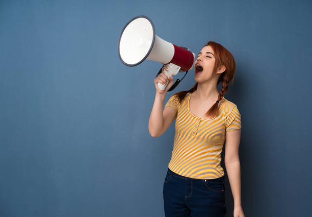 Young redhead woman over blue background shouting through a megaphone - Foto, Imagen