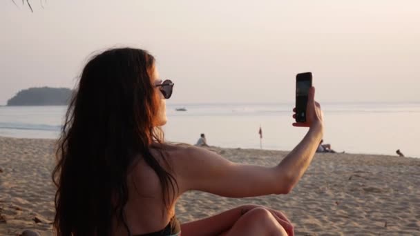 Young woman in a swimsuit and sunglasses takes pictures of golden sunset sea with phone sitting on the beach. Girl long hair holds hands with smartphone making photo memories of summer vacation with - Footage, Video