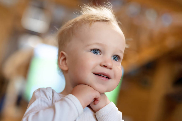 Cute caucasian blond toddler boy pleasantly surprised looking somewhere indoors. Adorable little child holding hands under chin and dreaming. Happy childhood concept - Fotografie, Obrázek